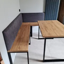 Kącik jadalny z drewnianym blatem stołu i siedziskiem ze ścianą obitą szarą tkaniną, na czarnych metalowych nogach, w nowoczesnym wnętrzu.