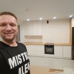 #Meble_Alex - Uśmiechnięty mężczyzna w czarnej koszulce z napisem na tle częściowo ukończonej, nowoczesnej kuchni z białymi szafkami i drewnianym blatem oraz czarną lodówką.