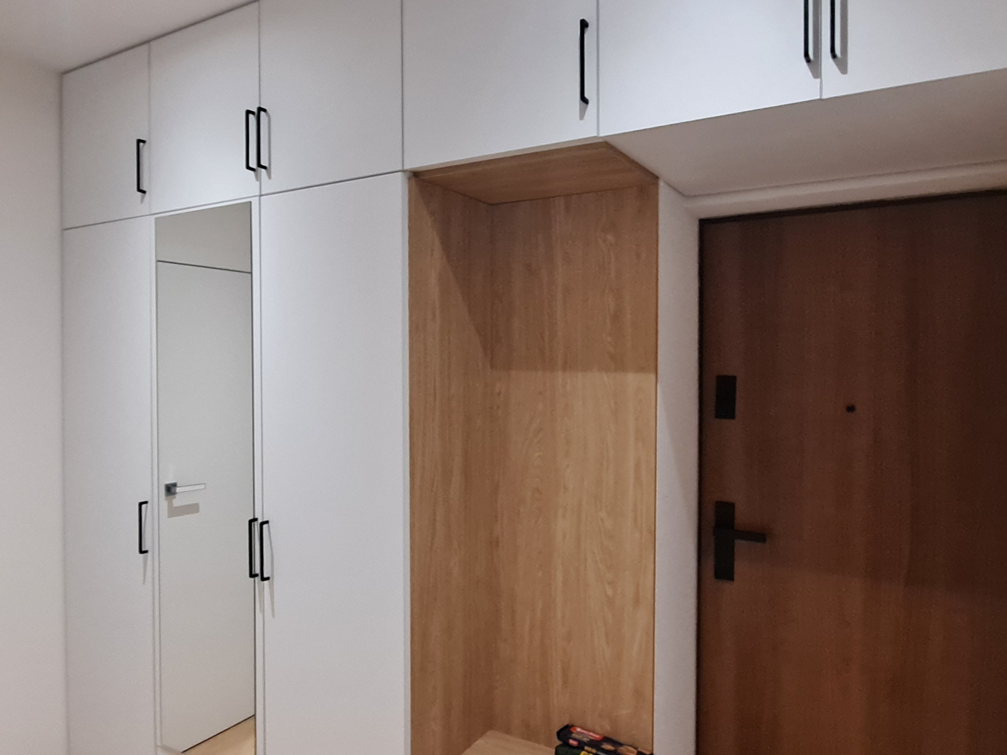 Biała szafa w przedpokoju z czarnymi uchwytami, obok drewniana wnęka i brązowe drzwi wejściowe.