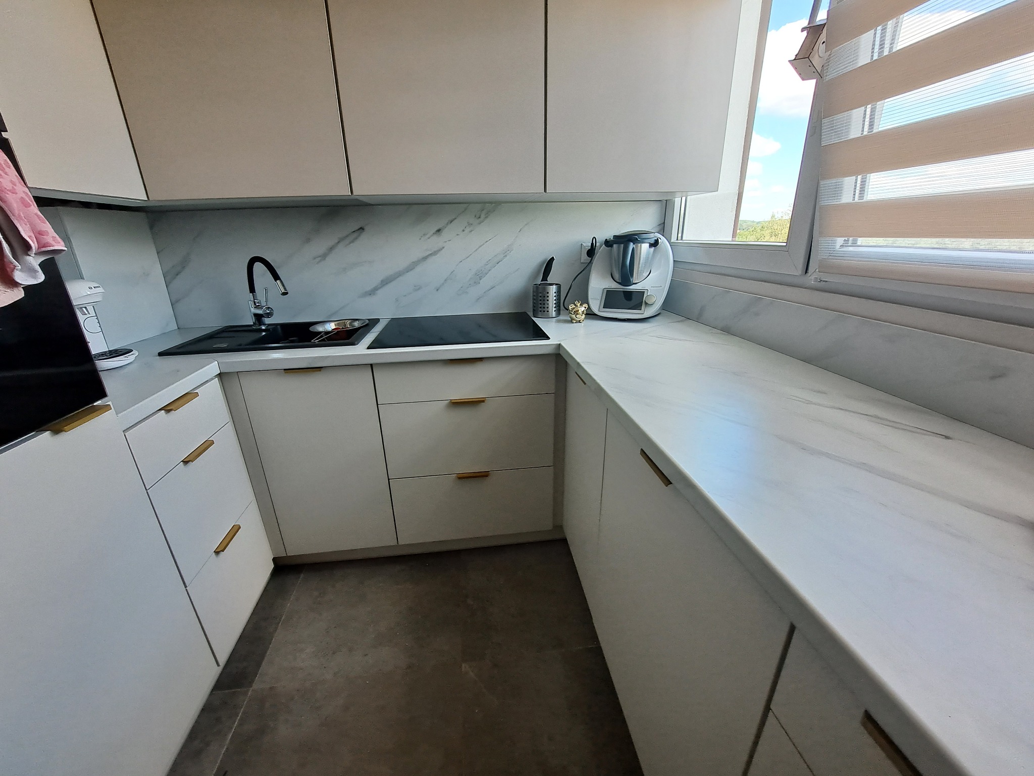 Jasna kuchnia z białymi szafkami, blatem imitującym marmur i złotymi uchwytami, widziana pod kątem, z widokiem na okno z żaluzjami.