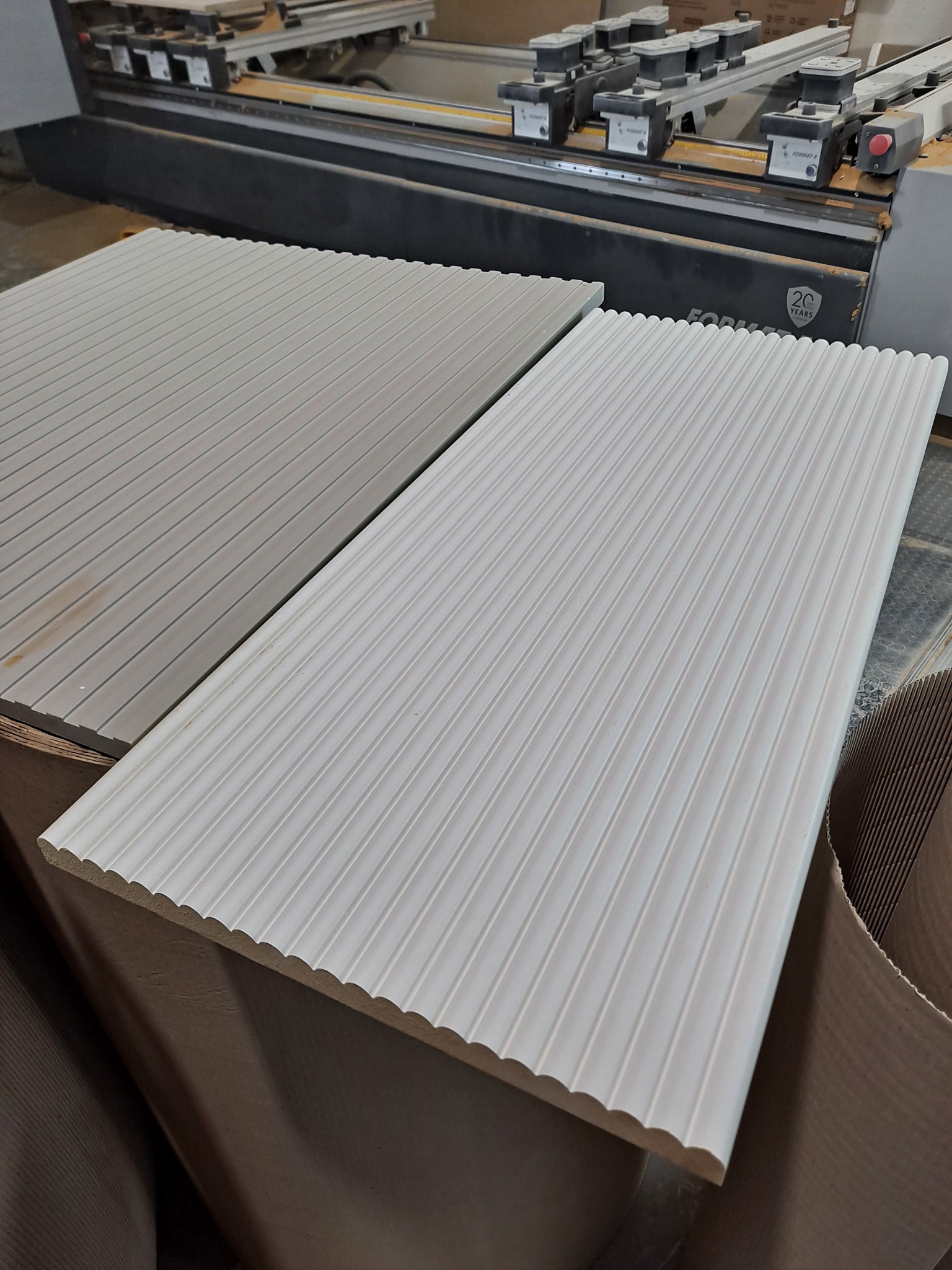 Dwa panele drewniane z frezowanymi rowkami, jeden szary i jeden biały, leżące na stosie materiałów, w tle widoczna maszyna CNC.