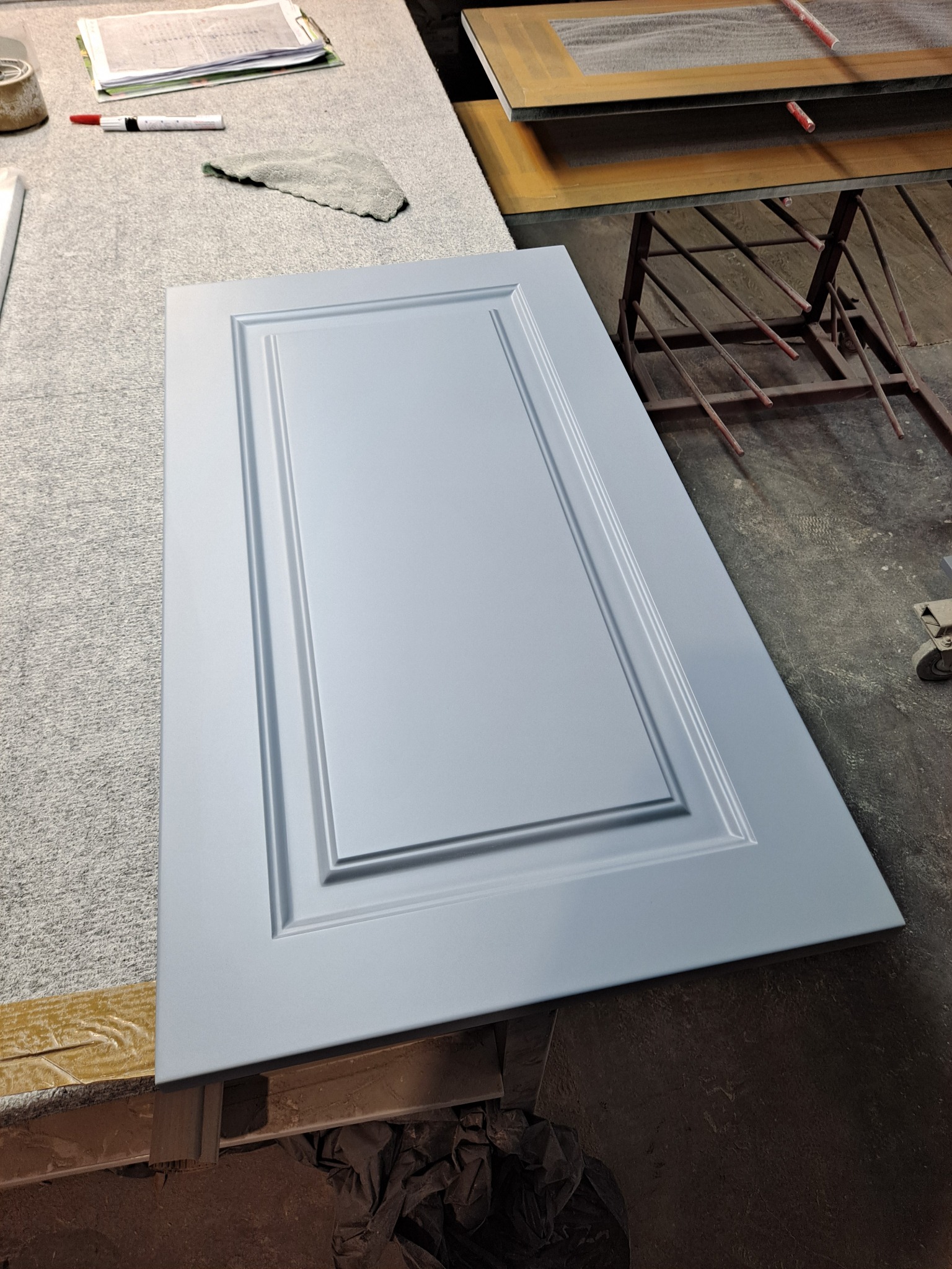 Pomalowane na jasnoniebiesko drzwiczki szafki z frezowanymi ramkami leżą na warsztatowym stole, obok widać dokumenty, szmatkę i inne elementy wyposażenia warsztatu stolarskiego.