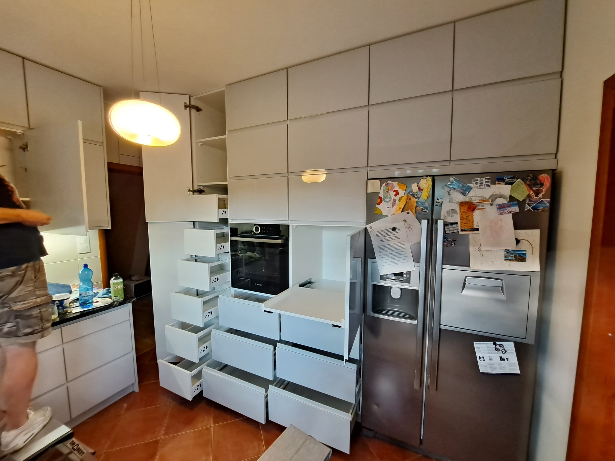 Montaż białej kuchni na wymiar z otwartymi szufladami, widoczny piekarnik i lodówka z magnesami, osoba na drabince montuje górne szafki