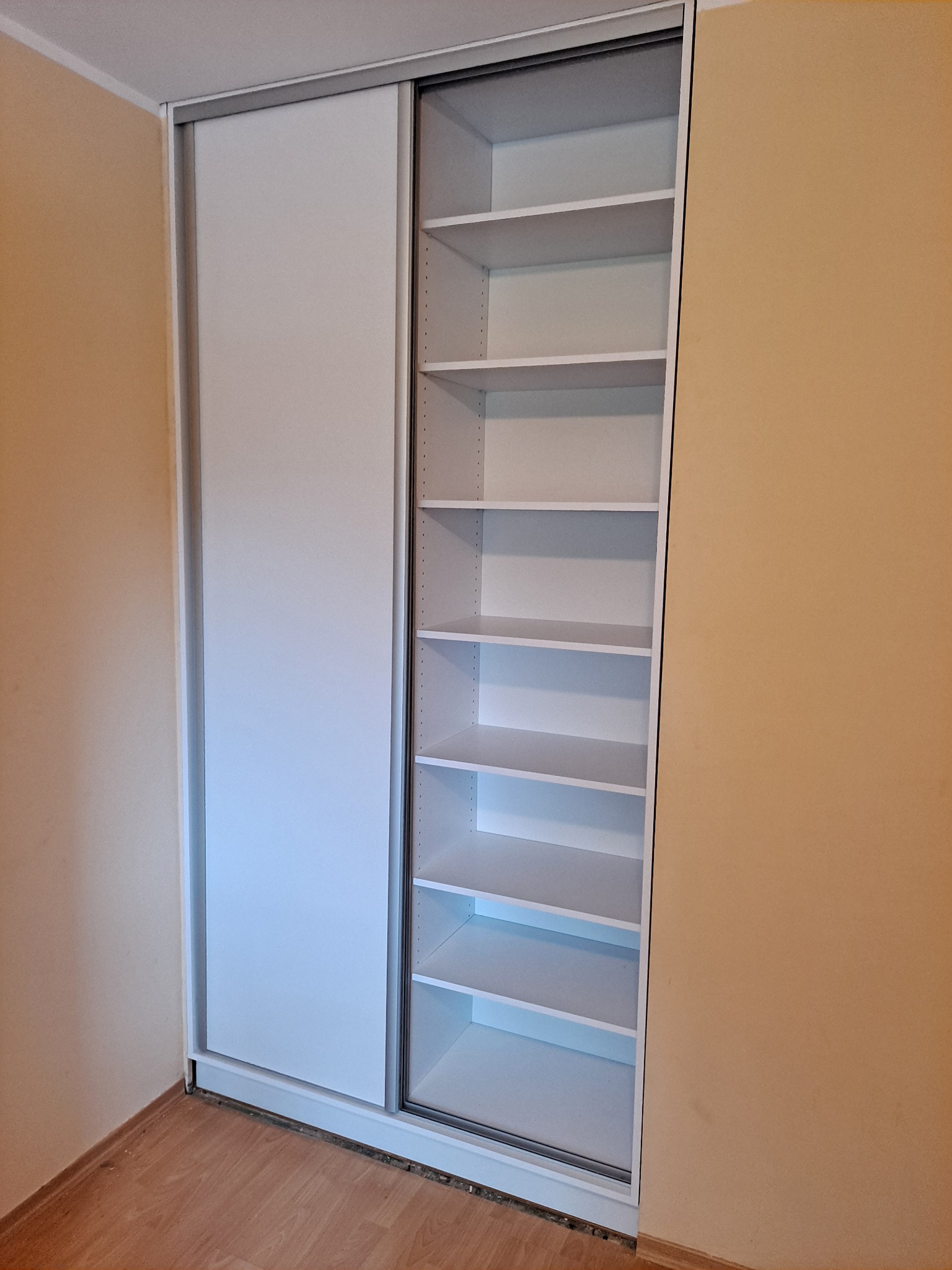 Biała szafa wnękowa z przesuwnymi drzwiami, widoczne puste półki, zamontowana w pomieszczeniu z beżowymi ścianami.