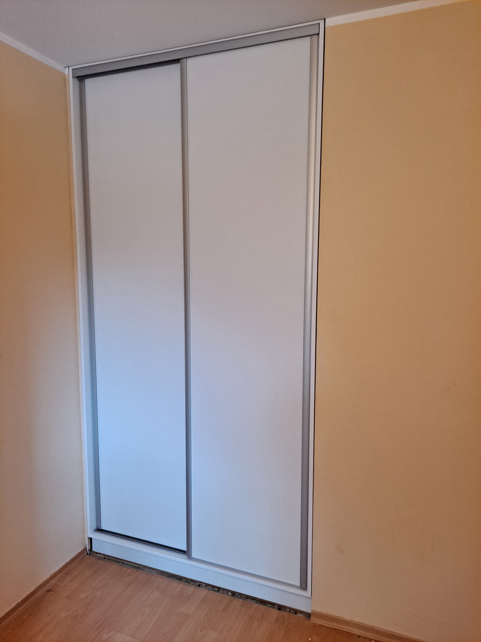 Biała szafa wnękowa z przesuwnymi drzwiami i srebrnymi obramowaniami, ustawiona na panelach podłogowych przy beżowej ścianie.