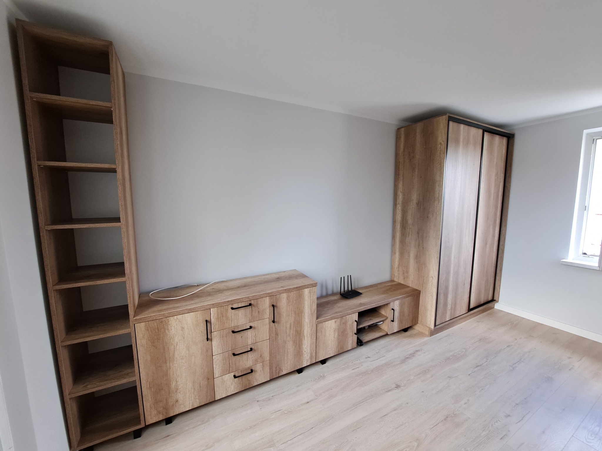 Zestaw mebli do salonu w odcieniach drewna, składający się z szafy przesuwnej, szafki RTV i regału, na podłodze panele w jasnym odcieniu drewna.