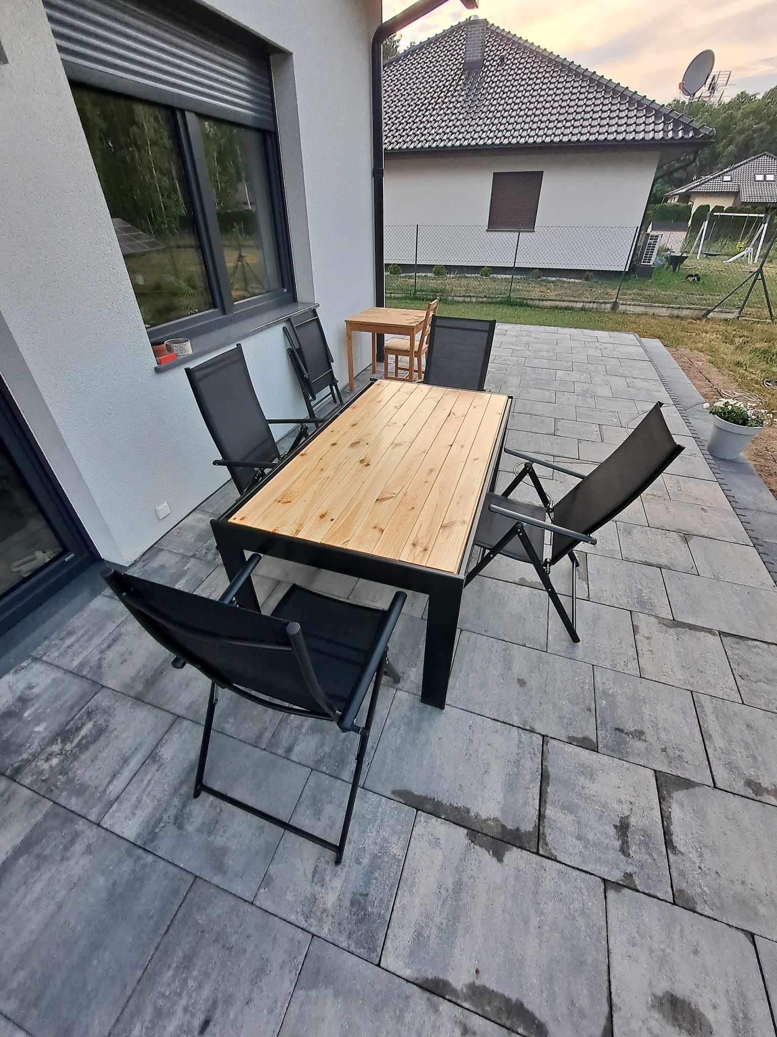 Zewnętrzny stół z blatem z jasnego drewna i czarnymi metalowymi nogami, otoczony składanymi krzesłami na szarym brukowanym tarasie przy domu z szarą elewacją.