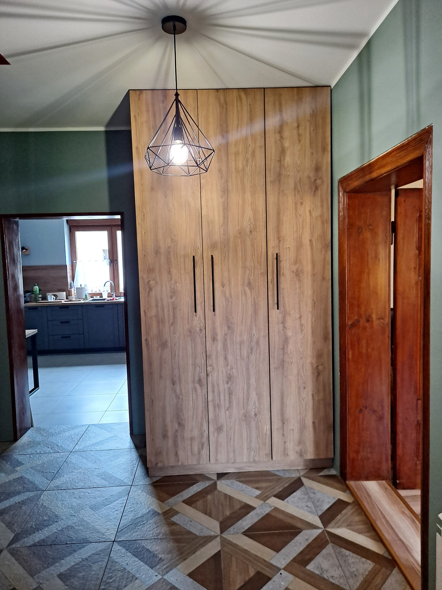 Trzydrzwiowa szafa z drewnopodobnym fornirem i czarnymi, pionowymi uchwytami, oświetlona wiszącą lampą z geometrycznym kloszem, w tle widoczny fragment kuchni.
