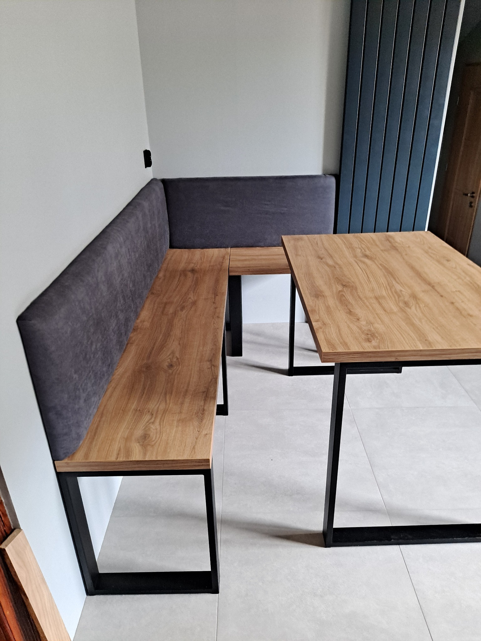 Kącik jadalny z drewnianym blatem stołu i siedziskiem ze ścianą obitą szarą tkaniną, na czarnych metalowych nogach, w nowoczesnym wnętrzu.