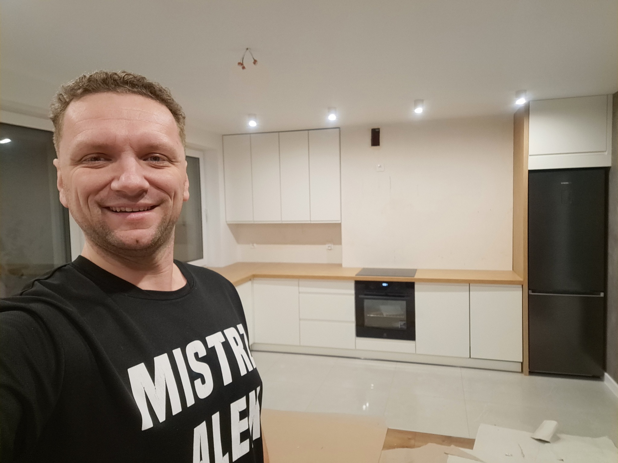 Uśmiechnięty mężczyzna w czarnej koszulce z napisem na tle częściowo ukończonej, nowoczesnej kuchni z białymi szafkami i drewnianym blatem oraz czarną lodówką.