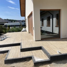 Damian Grzesiak - Układanie tarasu z płytek imitujących drewno, z czarnymi obrzeżami schodków, na tle nowoczesnego domu z dużym oknem i roletami zewnętrznymi. Widoczne krzyżyki dystansowe i biały kubek na schodku.