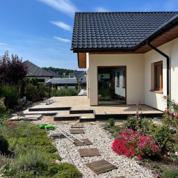 Damian Grzesiak - Tarasy z płytek ceramicznych w trakcie budowy, widoczne narzędzia i materiały budowlane, ścieżka z drewnianych płyt na żwirowym podłożu, ogród z kwiatami i krzewami, nowoczesny dom z ciemną...