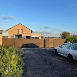 Nowoczesna posesja z przesuwną bramą wjazdową z poziomych paneli, szary samochód Skoda Octavia zaparkowany na żwirowym podjeździe, dom w tle w surowym stanie.