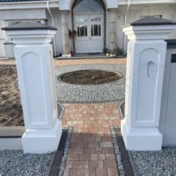 Widok na wejście do domu przez dwa białe filary, z wybrukowaną ścieżką z czerwonej kostki prowadzącą do okrągłego klombu z szarego kamienia, w tle biały dom z ciemnym dachem.