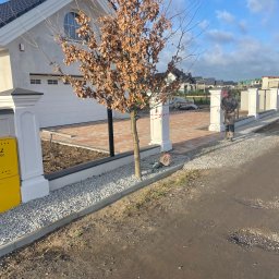 Damian Grzesiak - Trwa układanie czerwono-brązowej kostki na podjeździe w Kliniskach Wielkich, widoczny pracownik w odzieży roboczej, białe słupki ogrodzeniowe i podsypka z drobnego kamienia.