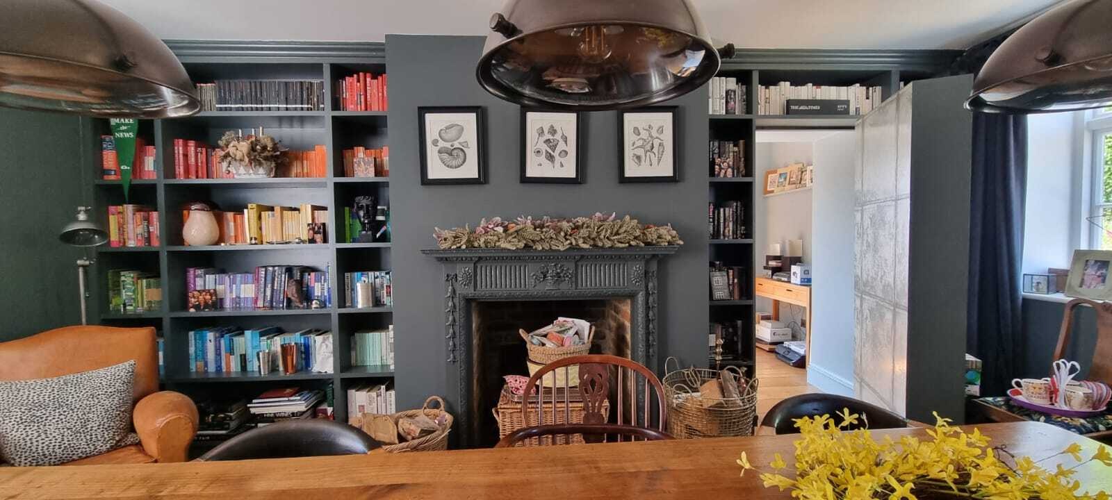 Szare wnętrze salonu z regałami na książki, kominkiem ozdobionym suszem, dużymi lampami wiszącymi i drewnianym stołem z żółtymi kwiatami.