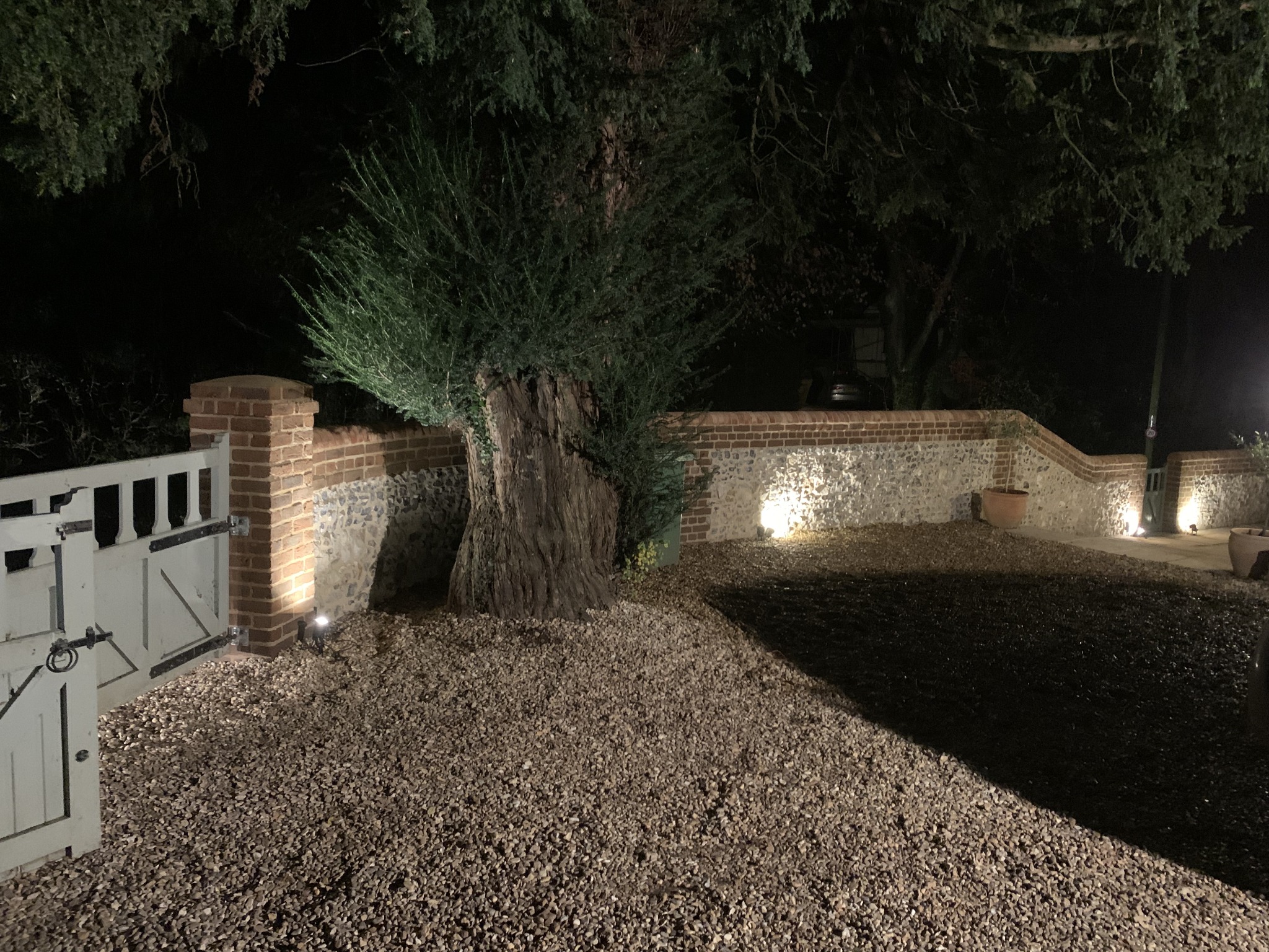 Nocne ujęcie na posesję z brukowanym podjazdem, oświetlonym niskimi lampami, murkiem z cegły i kamienia oraz drewnianą bramą wjazdową.