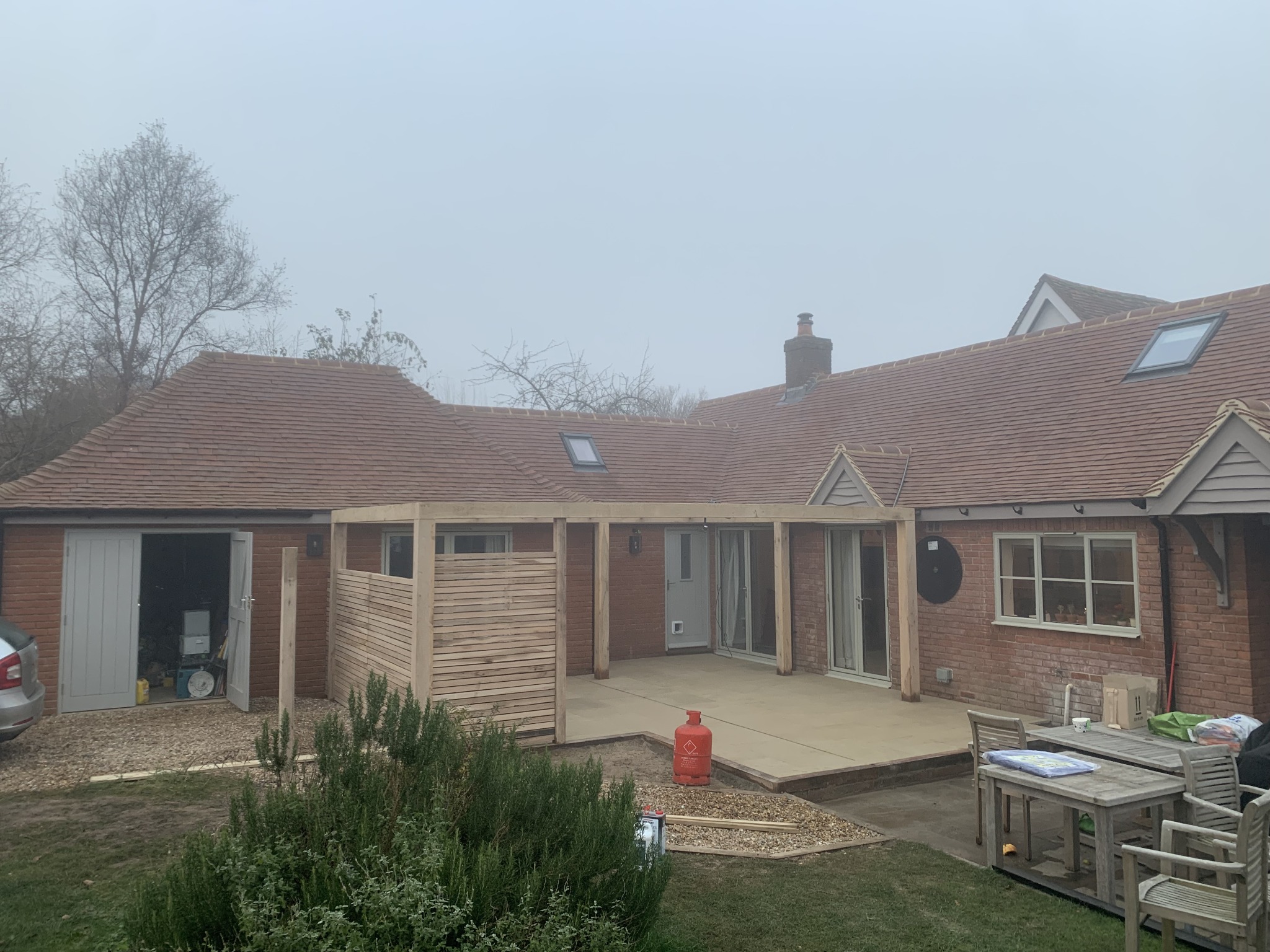 Budowa drewnianej pergoli przy domu z cegły, widoczna konstrukcja szkieletowa i częściowo zamontowane panele, na pierwszym planie butla gazowa i narzędzia, pochmurna pogoda.