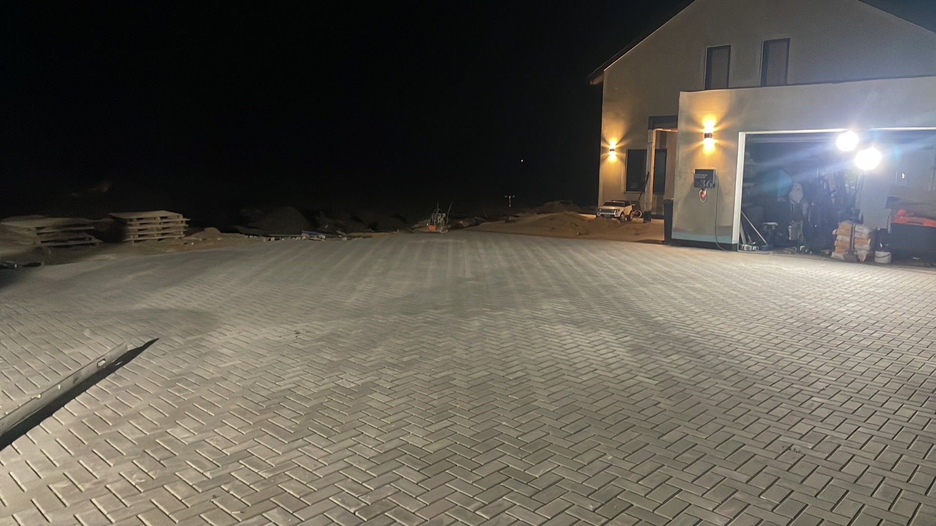 Nowo ułożona szara kostka brukowa w jodełkę na podjeździe przed domem z otwartym garażem w nocy, widoczne palety i maszyna brukarska.