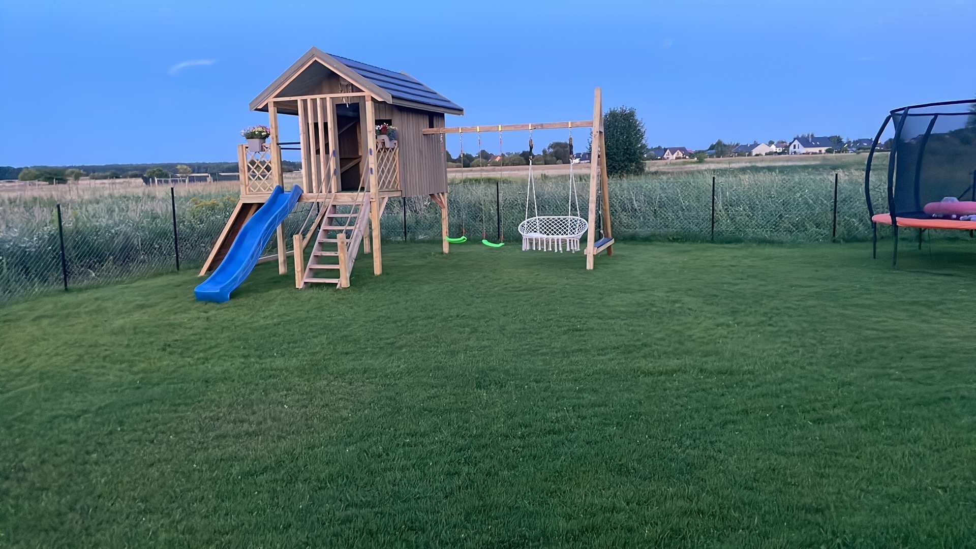Drewniany plac zabaw z domkiem, zjeżdżalnią, huśtawkami i trampoliną na trawniku w ogrodzie, widok na wieś w tle, wieczorny zmierzch.