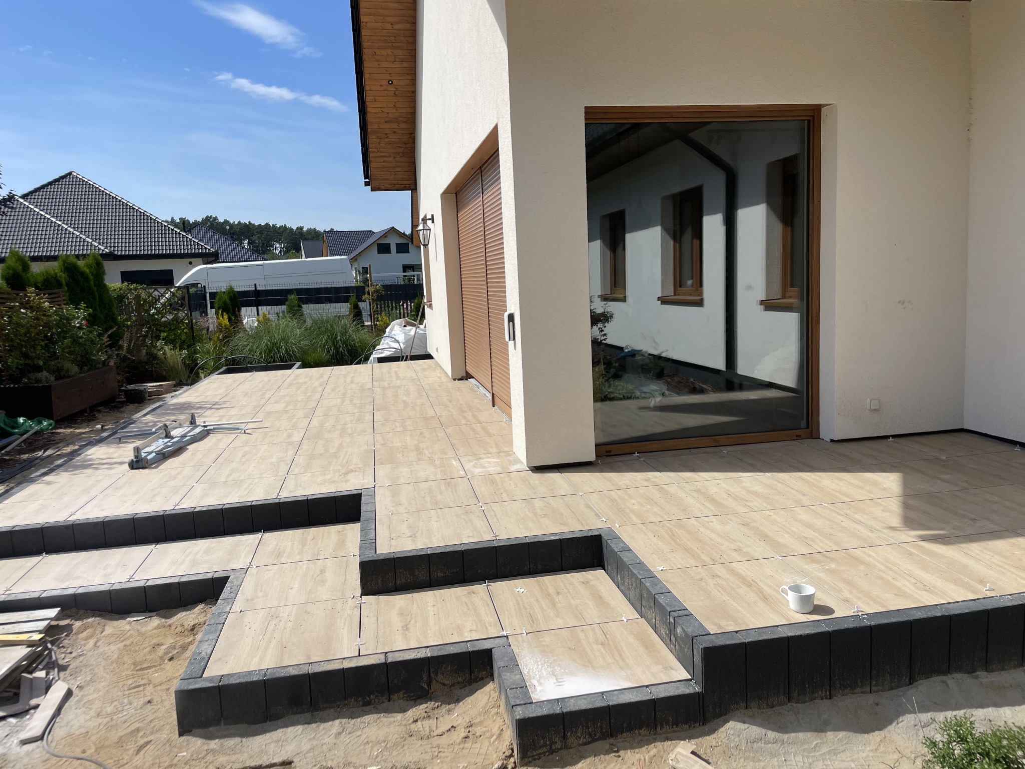 Układanie tarasu z płytek imitujących drewno, z czarnymi obrzeżami schodków, na tle nowoczesnego domu z dużym oknem i roletami zewnętrznymi. Widoczne krzyżyki dystansowe i biały kubek na schodku.