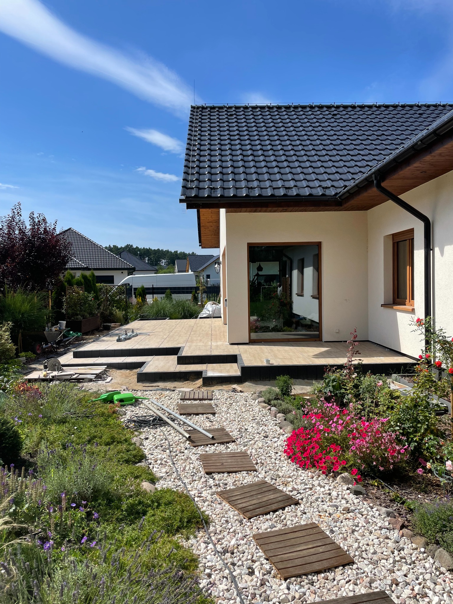 Tarasy z płytek ceramicznych w trakcie budowy, widoczne narzędzia i materiały budowlane, ścieżka z drewnianych płyt na żwirowym podłożu, ogród z kwiatami i krzewami, nowoczesny dom z ciemną...