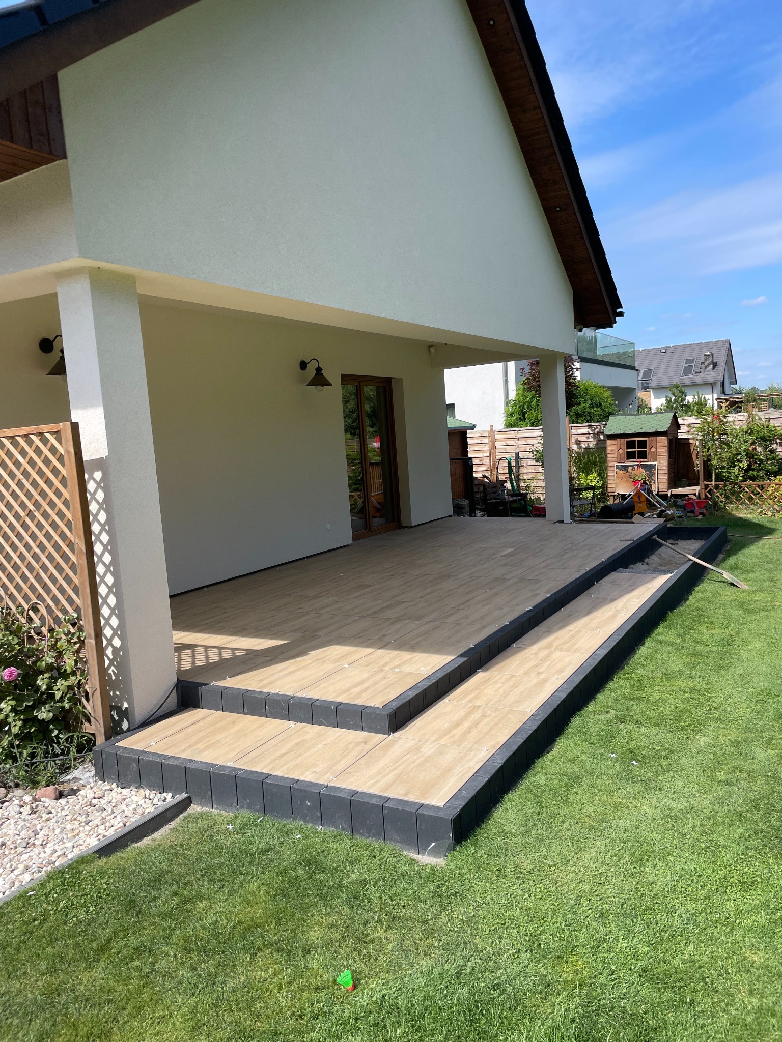 Nowoczesny taras z płytek imitujących drewno, z dwoma stopniami obłożonymi czarną kostką, przylegający do budynku z jasną elewacją i kolumnami, widok na zadbany trawnik i elementy ogrodu.