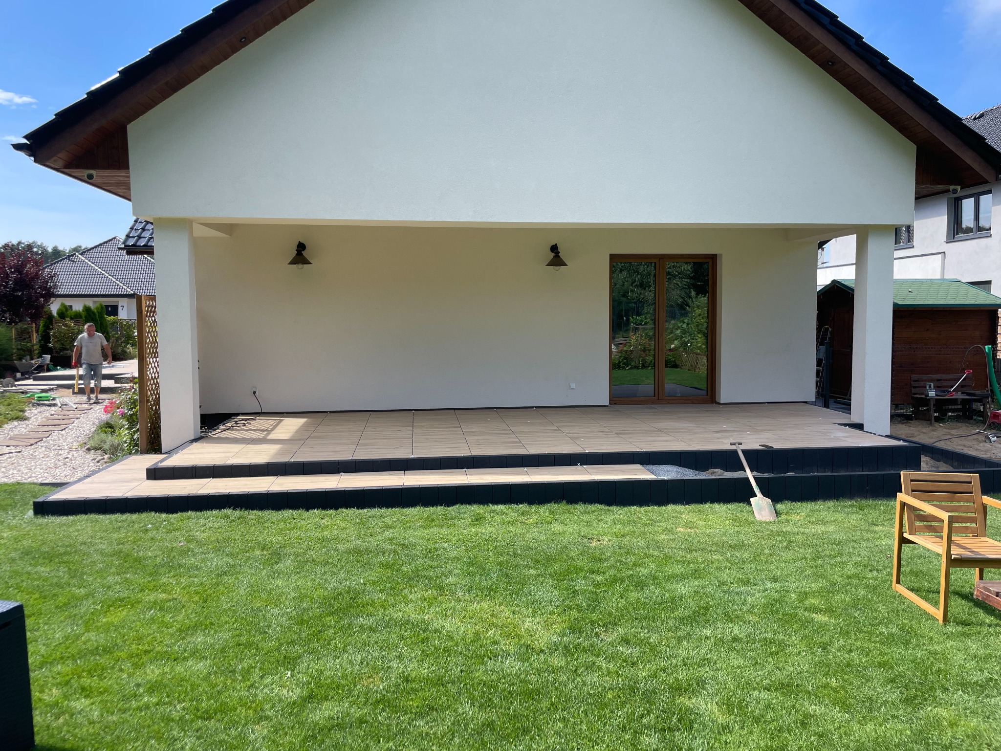 Nowoczesny taras z jasnych płytek ceramicznych, wykończony czarnym obrzeżem, w trakcie budowy, widoczna łopata i krzesło ogrodowe na trawniku, biała elewacja domu z oknem tarasowym w tle.