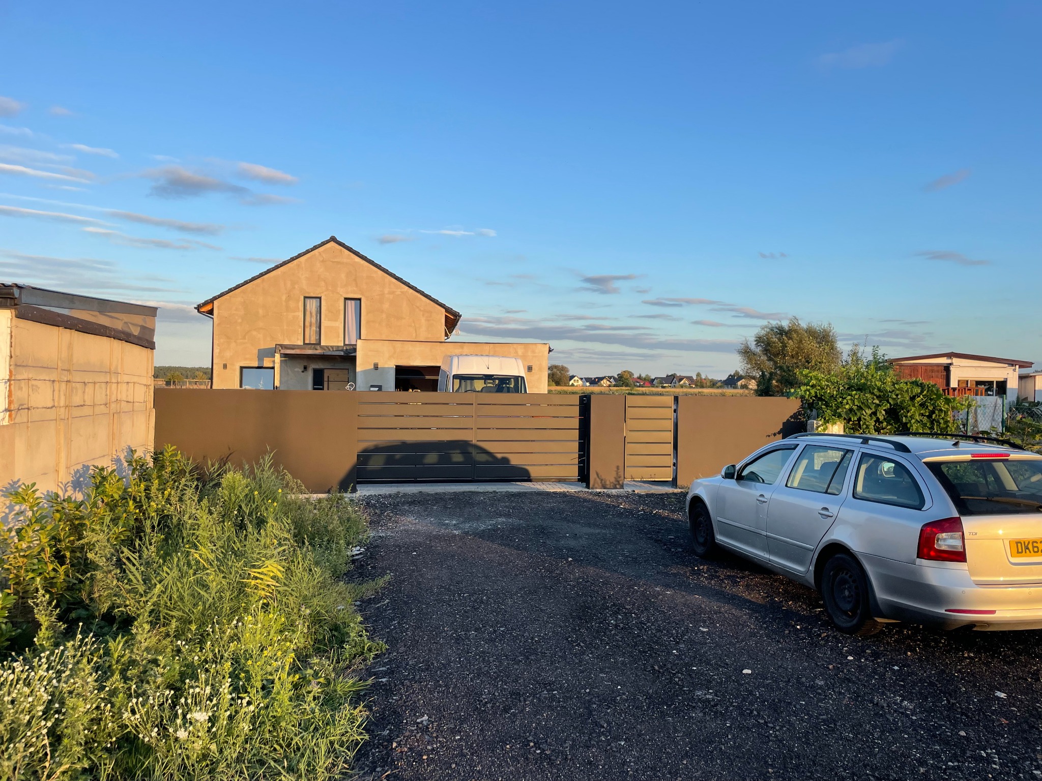 Nowoczesna posesja z przesuwną bramą wjazdową z poziomych paneli, szary samochód Skoda Octavia zaparkowany na żwirowym podjeździe, dom w tle w surowym stanie.