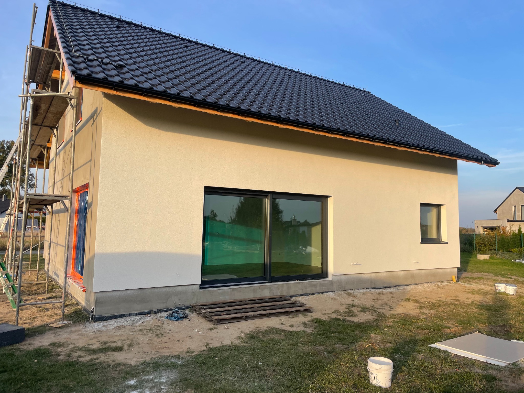 Dom jednorodzinny w trakcie prac wykończeniowych elewacji, widoczne rusztowania, okna zabezpieczone folią, czarna dachówka, ślady prac budowlanych na trawniku.