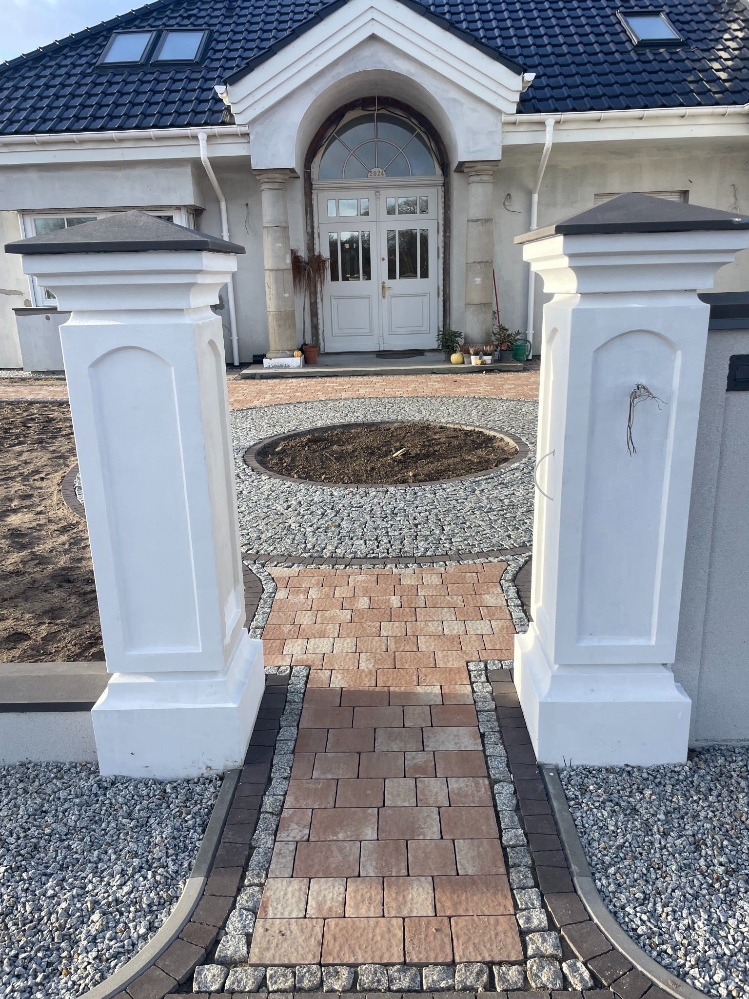 Widok na wejście do domu przez dwa białe filary, z wybrukowaną ścieżką z czerwonej kostki prowadzącą do okrągłego klombu z szarego kamienia, w tle biały dom z ciemnym dachem.
