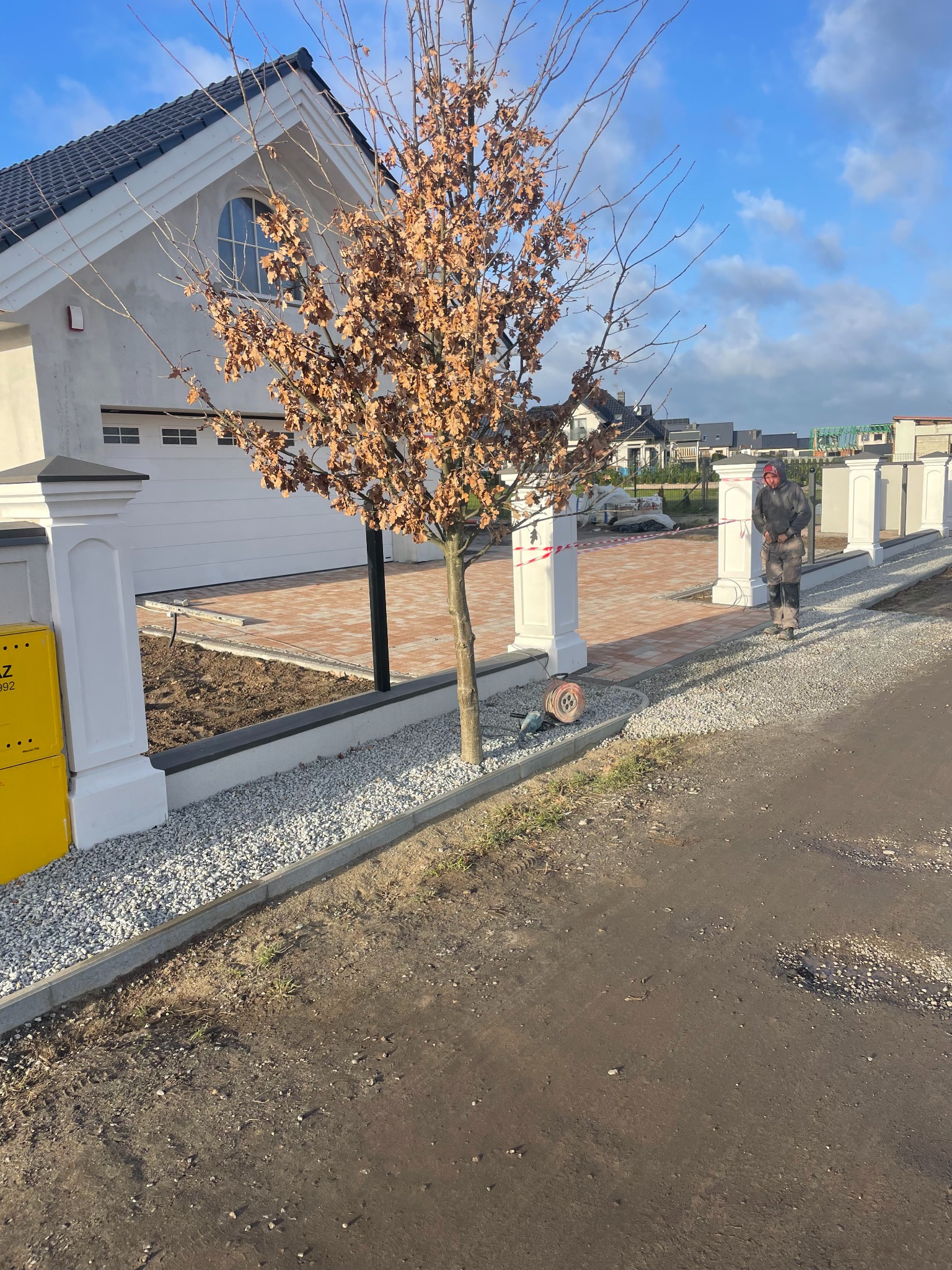 Trwa układanie czerwono-brązowej kostki na podjeździe w Kliniskach Wielkich, widoczny pracownik w odzieży roboczej, białe słupki ogrodzeniowe i podsypka z drobnego kamienia.