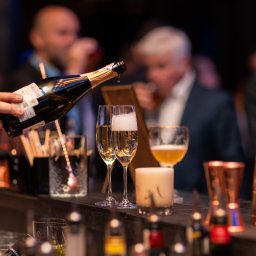 FotografNagrania.pl - Nalewanie szampana do kieliszków na barze, w tle rozmyte sylwetki osób, widoczne akcesoria barmańskie i świeca.