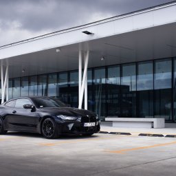 FotografNagrania.pl - Czarny samochód sportowy BMW M4 stoi na parkingu przed nowoczesnym budynkiem z przeszkloną fasadą i białym zadaszeniem, pochmurne niebo w tle.