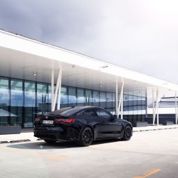 FotografNagrania.pl - Czarny samochód BMW M4 stoi na parkingu przed nowoczesnym budynkiem z szklaną fasadą i białymi kolumnami, widok z tyłu, pochmurne niebo.