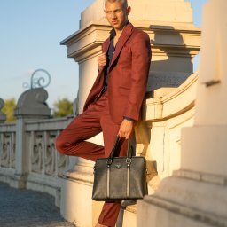 FotografNagrania.pl - Mężczyzna w bordowym garniturze i czarnych mokasynach, oparty o kamienną balustradę, trzyma czarną skórzaną torbę Prada.