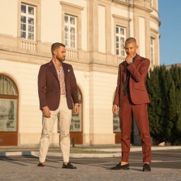 FotografNagrania.pl - Dwóch mężczyzn w eleganckich, bordowych garniturach pozuje na tle klasycznej architektury, jeden z koszulą w kwiatowy wzór, drugi z ręką pod brodą.