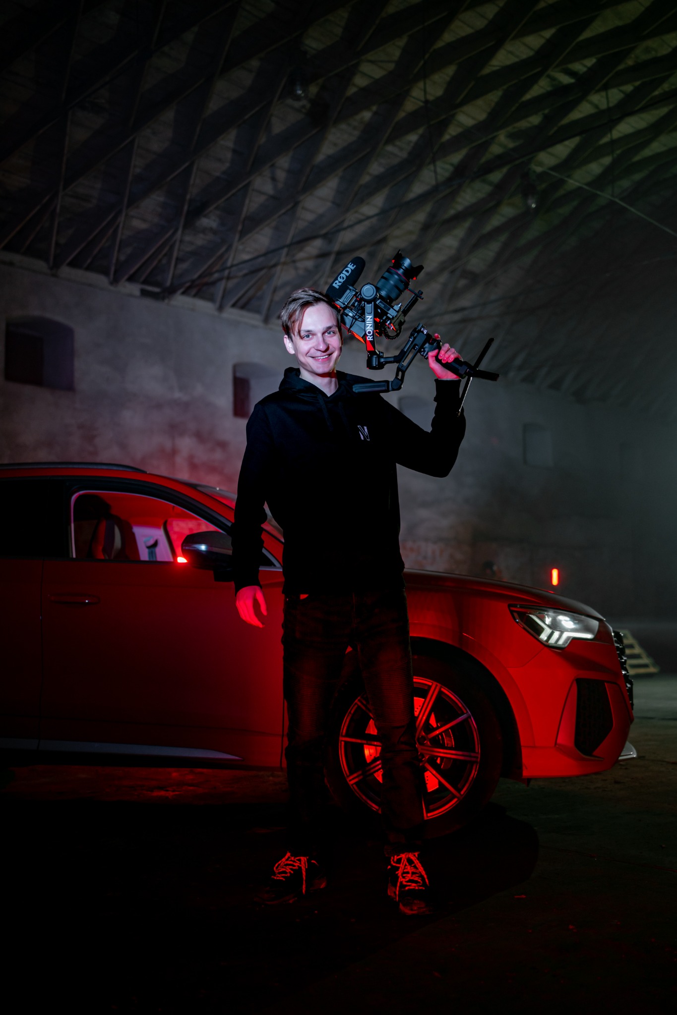 Uśmiechnięty mężczyzna w czarnej bluzie z kapturem trzyma kamerę z mikrofonami na stabilizatorze, w tle czerwony samochód i ceglane wnętrze budynku z widoczną konstrukcją dachu.