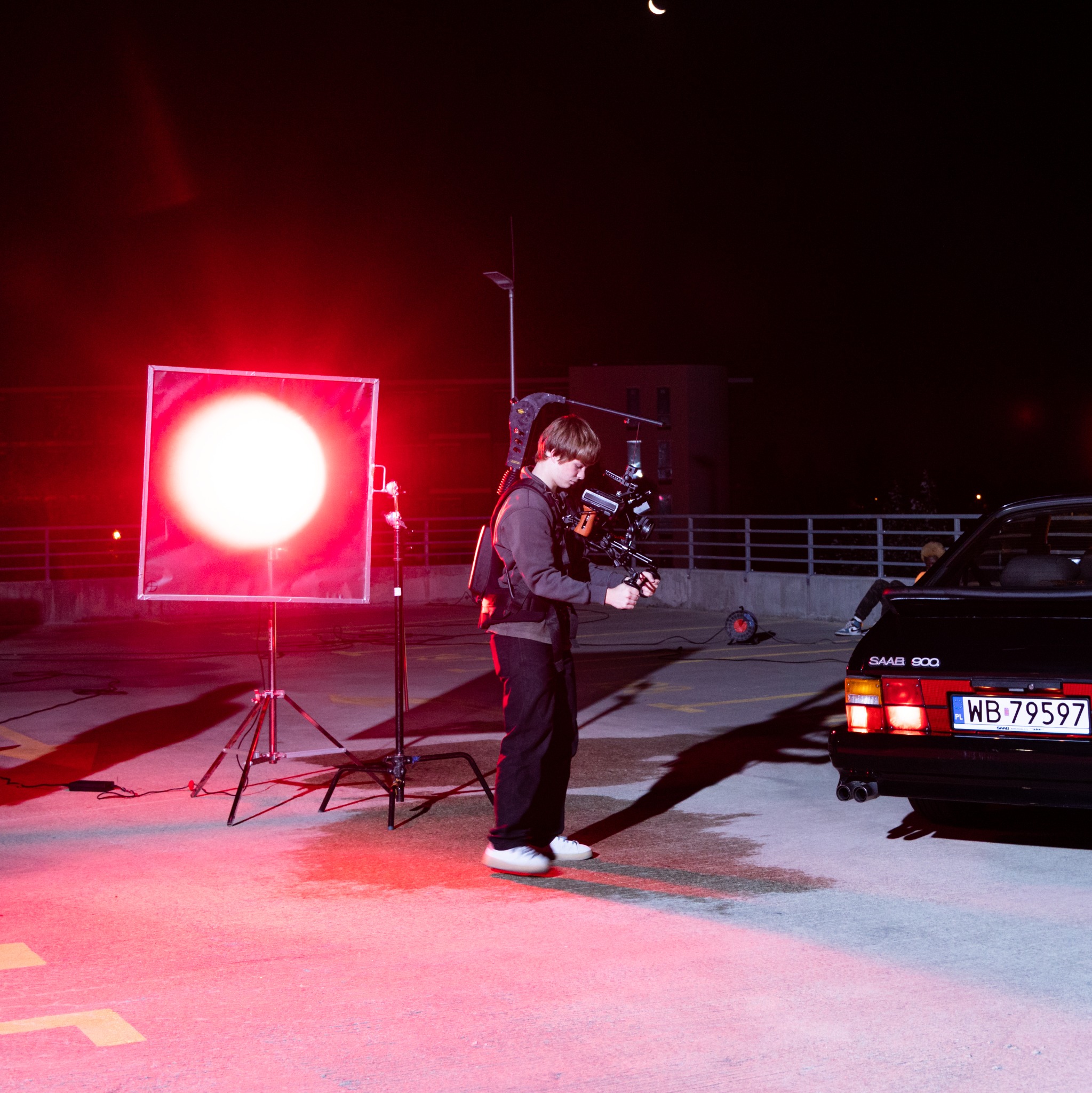 Kamerzysta z zamontowaną kamerą na stabilizatorze, oświetlony czerwonym światłem, filmuje samochód Saab 900 na parkingu nocą, widoczny sierp księżyca.