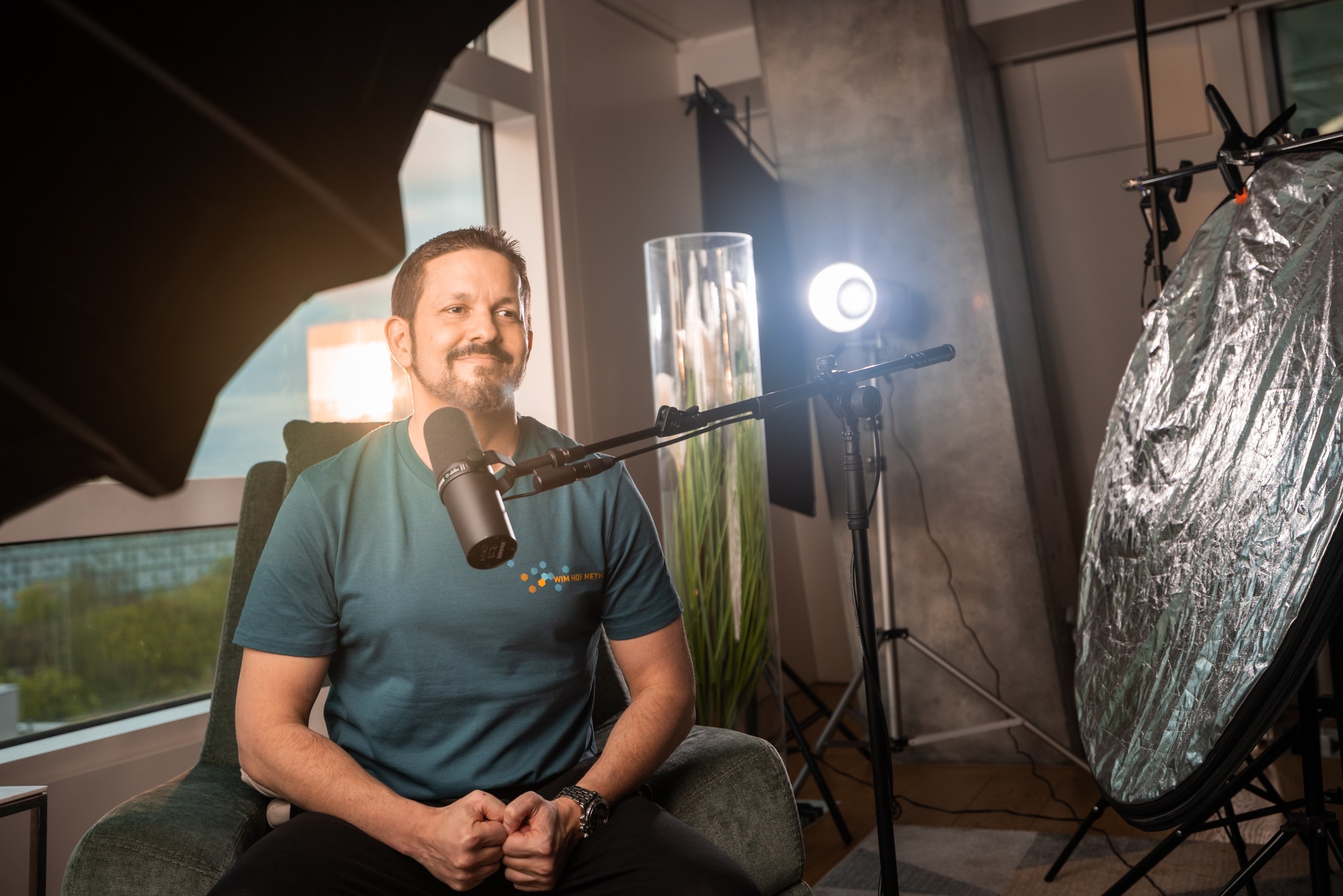 Mężczyzna z brodą siedzi w fotelu przed mikrofonem Shure SM7B, oświetlony lampami studyjnymi i reflektorem, okno w tle.