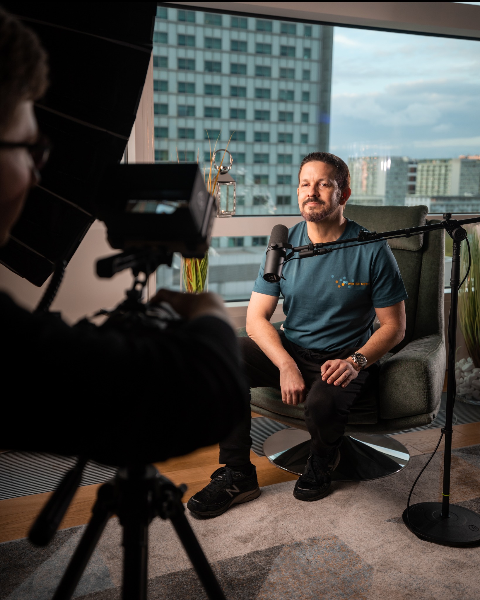 Mężczyzna w turkusowej koszulce siedzi na obrotowym fotelu podczas nagrywania w studio z mikrofonem Shure i softboxem; w tle okno z widokiem na miasto.