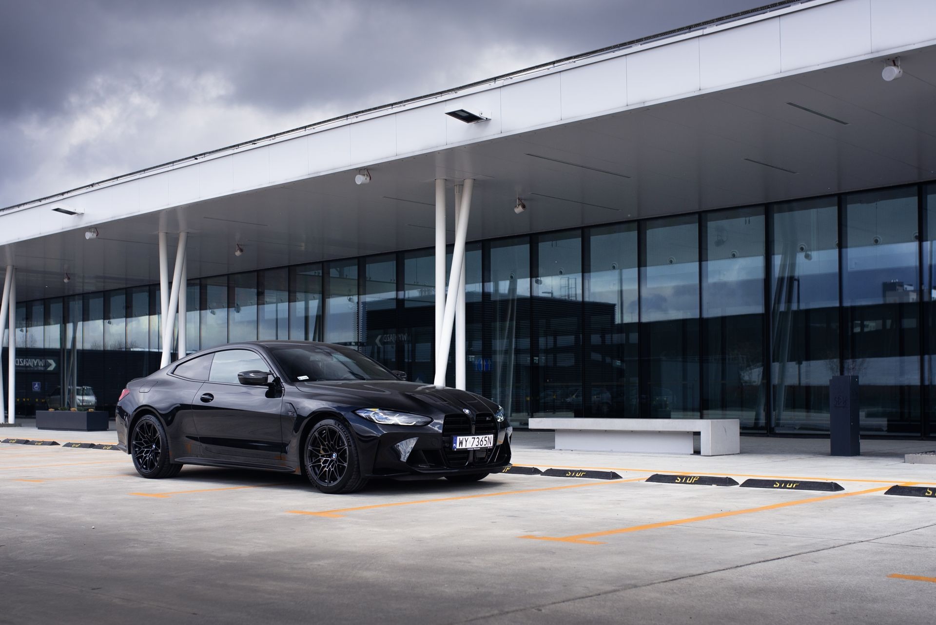 Czarny samochód sportowy BMW M4 stoi na parkingu przed nowoczesnym budynkiem z przeszkloną fasadą i białym zadaszeniem, pochmurne niebo w tle.