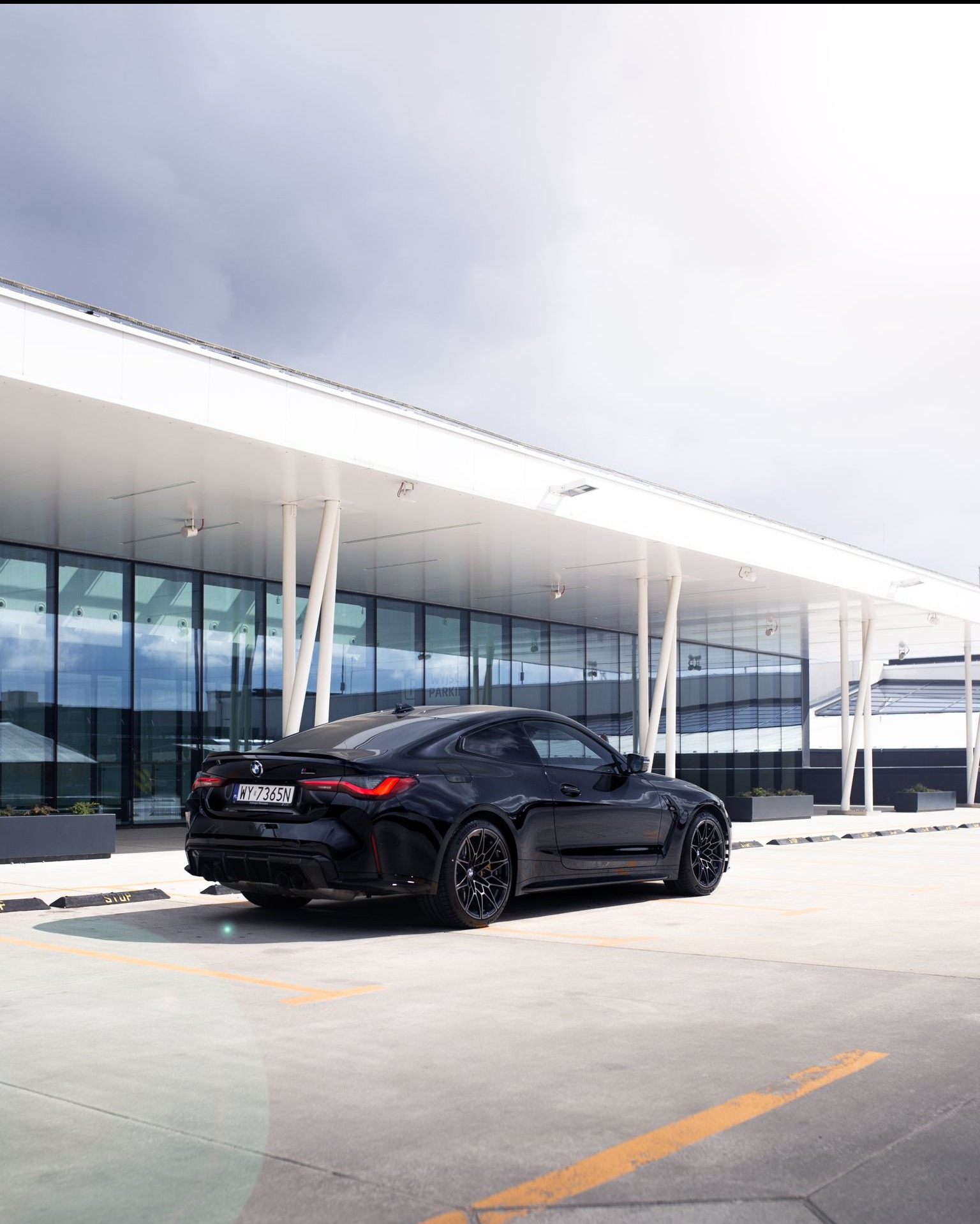 Czarny samochód BMW M4 stoi na parkingu przed nowoczesnym budynkiem z szklaną fasadą i białymi kolumnami, widok z tyłu, pochmurne niebo.