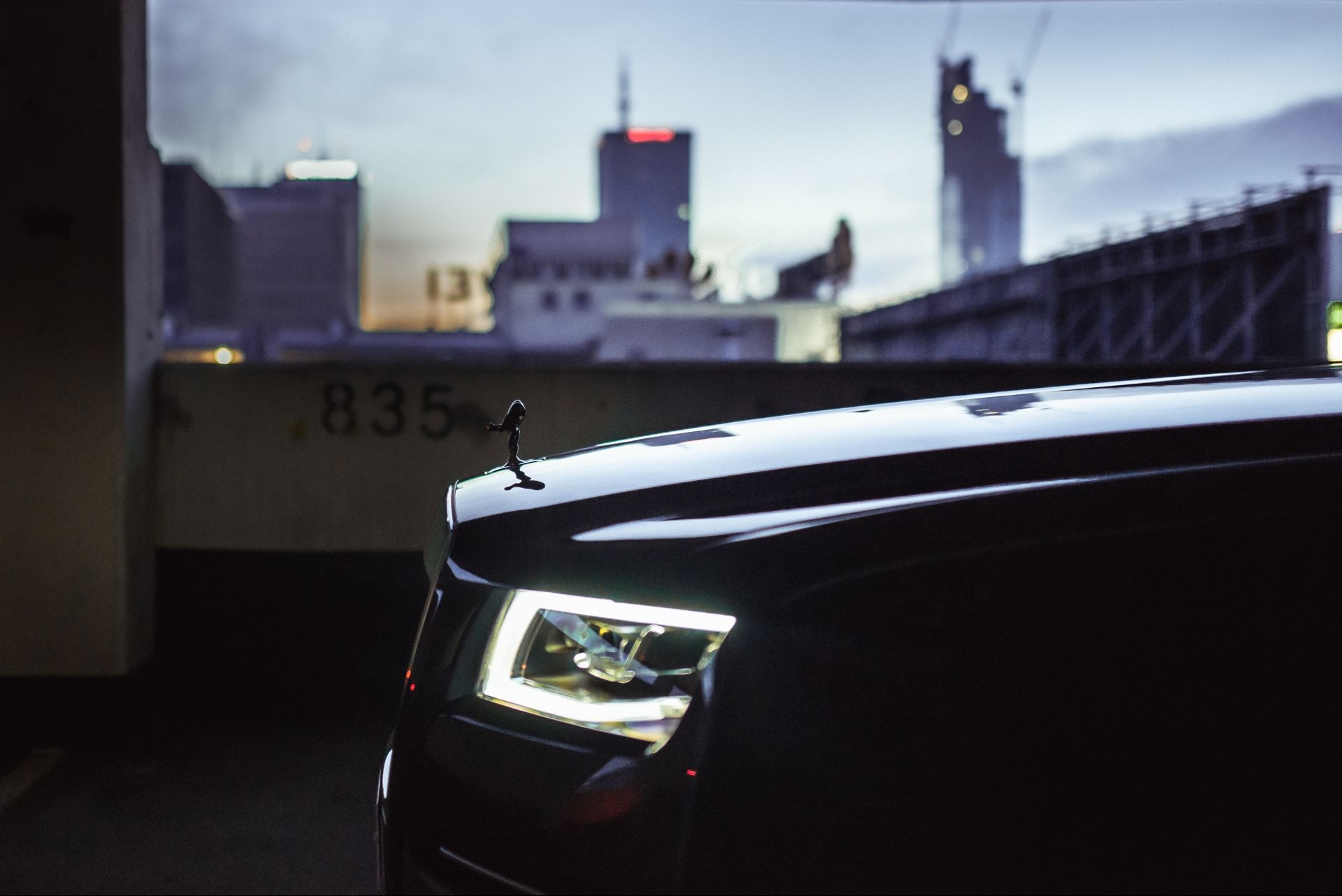 Fragment czarnego samochodu Rolls-Royce z podświetlonym reflektorem i figurką Spirit of Ecstasy na tle miejskiego krajobrazu z budynkami w budowie.