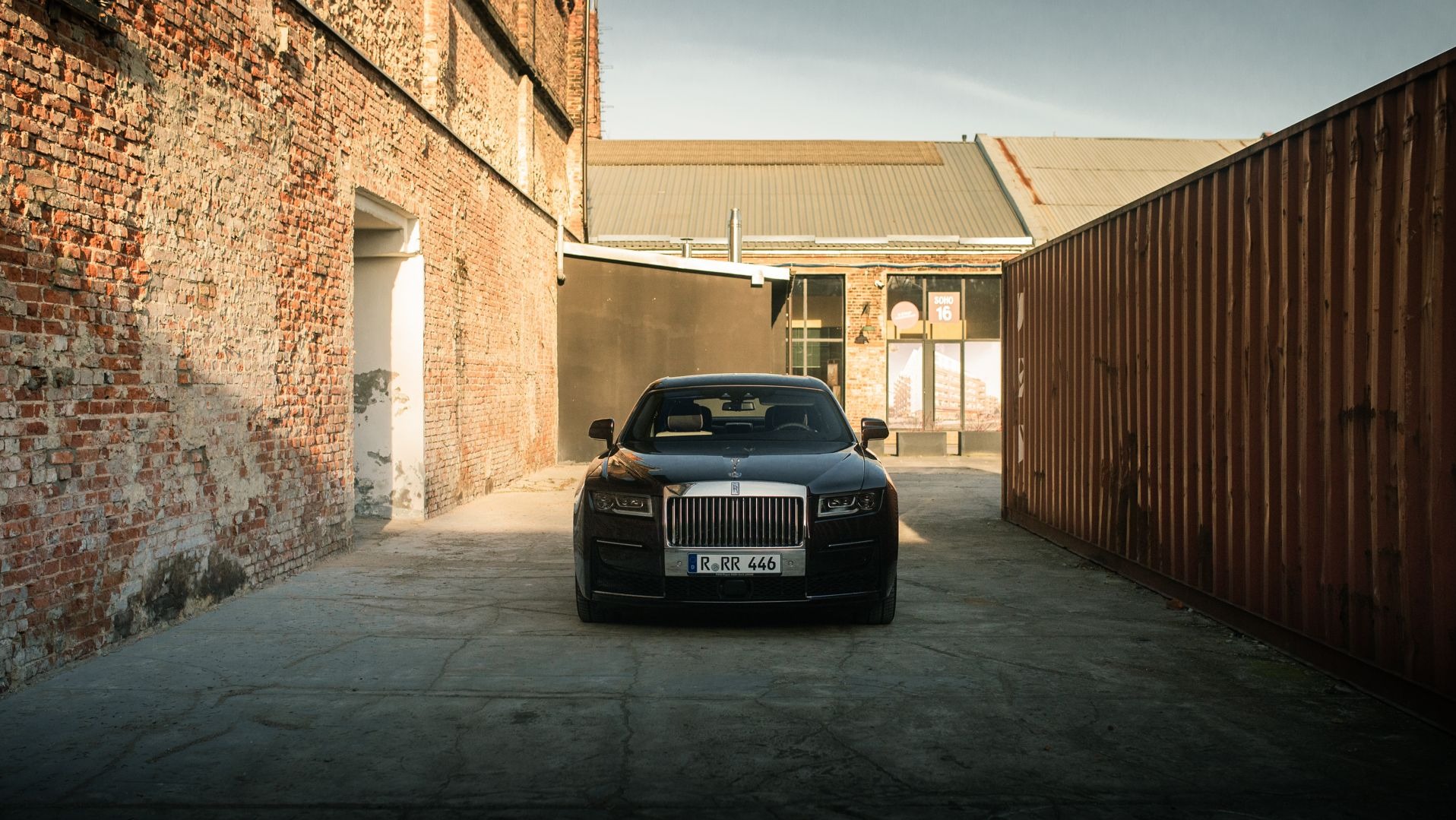 Czarny samochód marki Rolls Royce sfotografowany na zewnątrz, na tle ceglanej ściany i kontenera, w pochmurny dzień.
