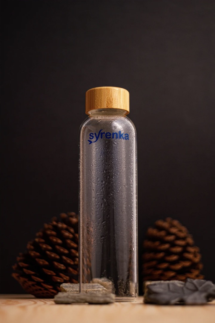 Szklana butelka z drewnianym korkiem, oznaczona niebieskim napisem 'Syrenka', otoczona szyszkami i kamieniami na drewnianym blacie, na ciemnym tle.