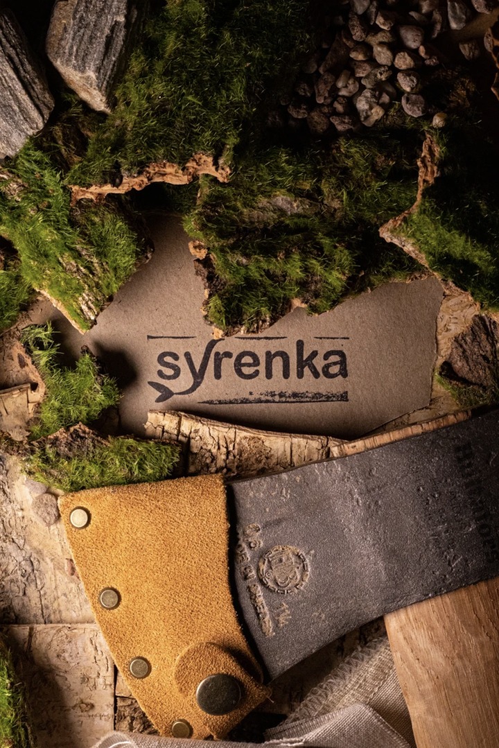 Toporek z brązowym skórzanym pokrowcem na tle kory drzew, mchu i logo 'syrenka' na brązowym papierze.