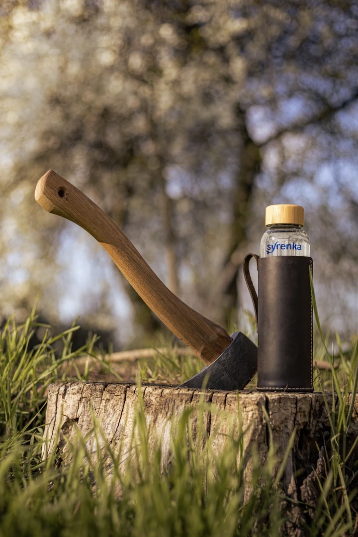 Toporek wbity w pień drzewa obok szklanej butelki z napisem Syrenka w skórzanym pokrowcu, na tle kwitnących drzew.