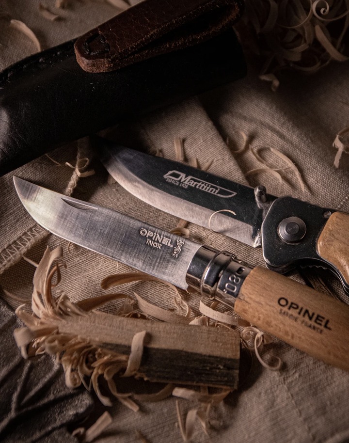 Dwa noże składane, Opinel i Marttiini, leżące na lnianym płótnie obok drewnianych wiórów i skórzanego futerału; ciepłe, boczne oświetlenie podkreśla fakturę materiałów.