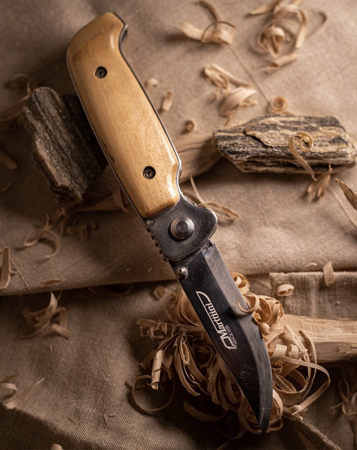 Składany nóż marki Marttiini z drewnianą rękojeścią, leżący na lnianym płótnie w otoczeniu wiórów drewna i fragmentów skał.